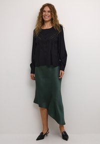 Blouse noire avec des détails en dentelle, associée à une jupe asymétrique en satin verte. Des chaussures noires à bout pointu complètent l'ensemble.