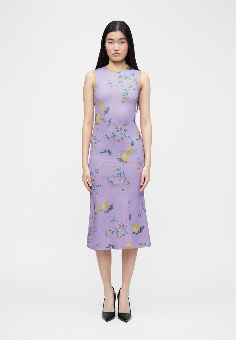 Νεαρή γυναίκα στέκεται φορώντας μίντι φόρεμα λιλά χωρίς μανίκια με λεμονί και λουλουδάτο μοτίβο και μαύρες μυτερές ψηλοτάκουνες γόβες μπροστά σε μονόχρωμο φόντο.