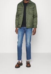 Chaqueta verde acolchada con dos bolsillos en el pecho, usada sobre una camiseta negra, combinada con jeans azul claro y mocasines marrones con suelas negras.