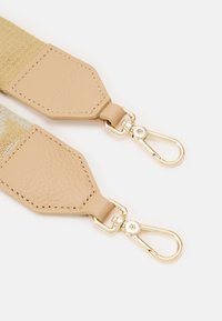 Dwa beżowe skórzane końce pasków z złotymi metalowymi obrotowymi karabińczykami przymocowanymi do materiałowej taśmy, leżące płasko na białym tle.