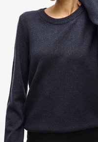 Personne portant un pull en maille bleu marine à manches longues et col rond côtelé, coupé à la taille, sur fond blanc.