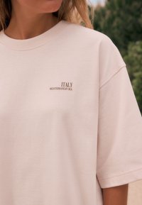 T-shirt oversize rose clair avec un col rond. Présente le texte "ITALY MEDITERRANEAN SEA" brodé en marron sur la poitrine. Texture douce.
