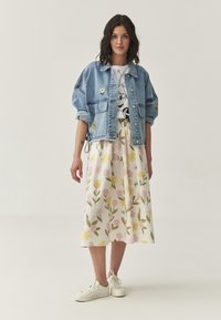 Lichte denimjas met geborduurde patches, wit graphic T-shirt, bloemen midi-rok in pastelkleuren, en witte sneakers.