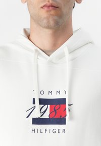 Weißer Hoodie mit Kordelzug und einem grafischen Druck, der "TOMMY", "1985" und "HILFIGER" in navyblauen und roten rechteckigen Akzenten zeigt. Glatte Textur.