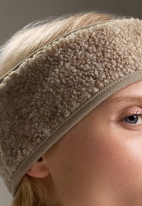Beige fleecový čelenka s texturovaným, nadýchaným povrchem a hladkým, přiléhavým páskem; detailní záběr odhalující měkkost a design látky.