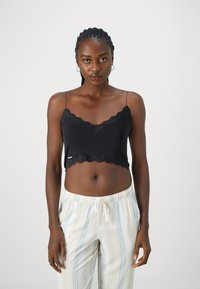 Top cropped preto com rendas, apresentando alças finas e bainha em forma de onda. Combinado com calças de pijama listradas claras.