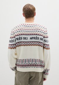 Cremefarbener Strickpullover mit geometrischen und Blattmustern in Rot, Blau und Senfgelb, mit einem Schriftband "APRÈS SKI" quer über der Brust.