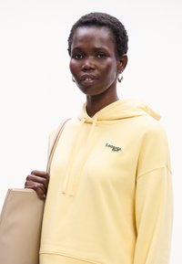 Felpa gialla in cotone con tasca a marsupio e cappuccio con cordino. Presenta un logo ricamato verde. Il modello tiene una borsa tote beige.