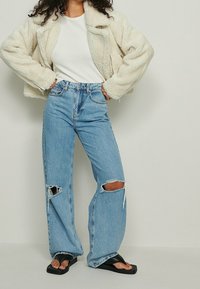 Chaqueta de piel sintética crema, top blanco de canalé, jeans de tiro alto en azul claro con desgarros en las rodillas, y sandalias planas negras.