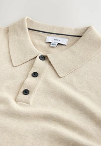 Béžová polo košile s texturovanou tkaninou, vybavená knoflíkovou légou na tři knoflíky a tmavými knoflíky, s kontrastním proužkem na límci. Viditelný štítek značky.