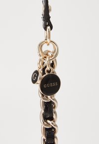 Must chain kuldse metallist lingidega, millel on kaks sildiga: üks ümmargune must silt, millel on kuldne tekst "GUESS" ja väiksem ringikujuline silt, millel on "G.".