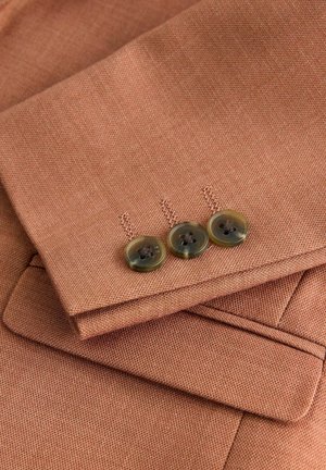 Braun strukturiertem Stoff mit drei grünlichen Knöpfen und detaillierter Naht am Ärmel eines Kleidungsstücks, das maßgeschneiderte Designelemente zeigt.