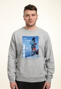 Uomo che indossa una felpa grigio chiaro con grafica di un alpinista, bussola e testo "Since 1981 Durham Mountaineering" sul davanti.