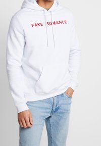 Vit hoodie med texten "FAKE ROMANCE" i röd, med känguruficka, justerbar huva och ribbstickade ärmar, ihop med ljusblå jeans.