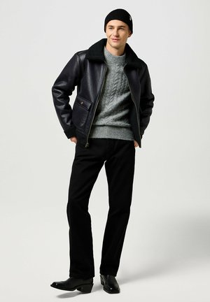 Leather bomber jacket with a shearling collar, gray knitted sweater, black trousers, and black heeled boots; neutral background.