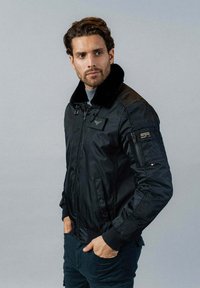 Veste bomber noire avec un col en fausse fourrure, poches zippées et un patch sur la poitrine. Texture lisse et design rembourré.
