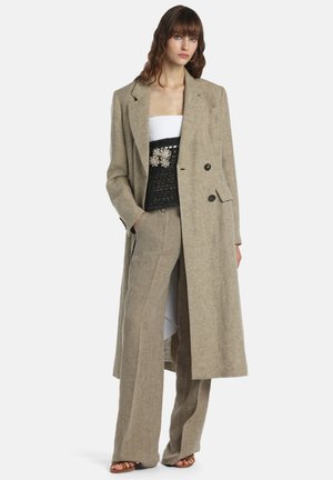 Kobieta ubrana w beżowy długi blezer i pasujące szerokie spodnie, z białą bluzką i czarnym ozdobnym gorsetem, stojąca z rękami w kieszeniach.