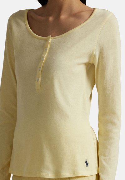 Femme portant un haut henley manches longues jaune pâle à quatre boutons avec un petit logo brodé près de l'ourlet.
