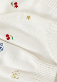Witte gebreide trui met geborduurde rode kersen en gouden sterren, met ribboorden en textuur. Inclusief blauwe gedetailleerde stiksels.