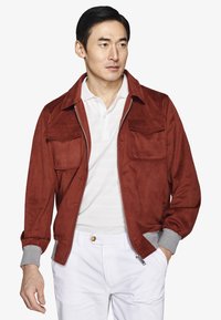 Homme portant une veste couleur rouille avec des poches devant, par-dessus un polo blanc et un pantalon blanc, regardant sur le côté avec une main dans la poche.