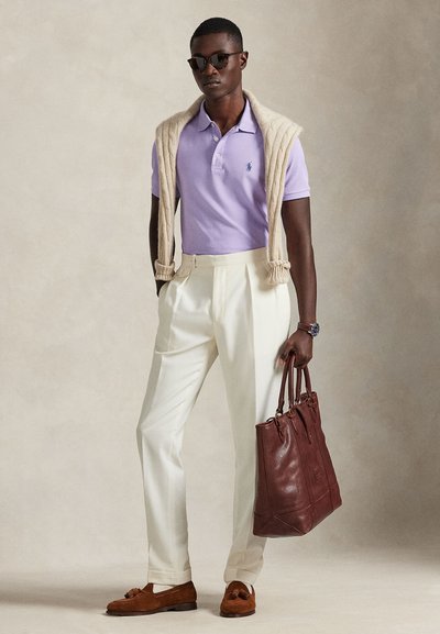 Ανοιχτό μωβ πόλο μπλουζάκι με κεντημένο λογότυπο, κρεμ παντελόνι με πιέτες, ταμπά σουέντ μοκασίνια και καφέ δερμάτινη τσάντα tote. Πλεκτό γιλέκο περασμένο πάνω από τους ώμους.