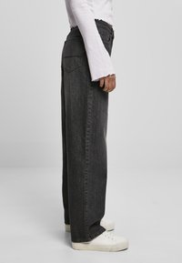 Vaqueros de mezclilla negros de tiro alto con corte holgado, bolsillos laterales y costuras visibles. Combinados con zapatillas blancas y una camiseta de manga larga.