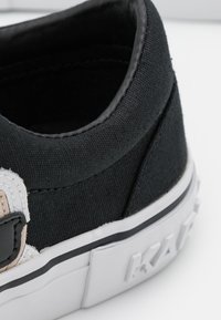 Gros plan sur le talon d'une basket en toile noire avec une semelle en caoutchouc blanche, présentant le nom de la marque embossé et un patch logo partiel.