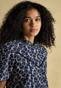 Tumedat sinine pluus valgete lillede tikandiga, volangkaelus ja pumbatud lühikesed varrukad; keeruline esikujundus ja pitsilised aksessuaarid.