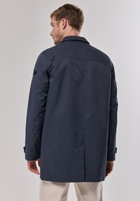 Abrigo de marinero con una silueta recta, mangas largas y cuello. Presenta detalles cosidos y una tela contrastante en la manga izquierda.