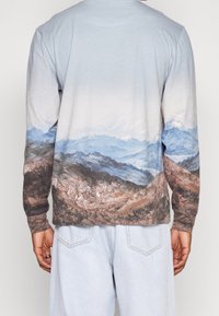 Langarmshirt mit Grafik eines Gebirgslandschaft. Hellblauer Hintergrund mit erdigen Tönen, strukturierter Stoff und lockere Passform.