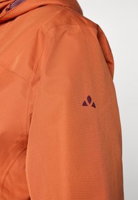 Artimas oranžinės striukės rankovės vaizdas, su tekstūruotu audiniu ir purpurine logotipu ant rankos, nurodantis prekės ženklo dizainą ir stilių.
