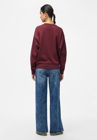 Mulher com cabelo trançado de costas, vestindo um moletom borgonha, jeans de perna larga azuis e sapatos castanhos, contra um fundo branco.