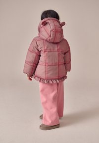 Giacca trapuntata rosa con motivo a quadri e orlo arricciato; abbinata a pantaloni rosa a gamba larga e scarpe slip-on marroni. Cappuccio con orecchie.