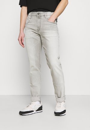 Personne portant un jean gris clair retroussé aux chevilles, des baskets blanches et noires, avec une main dans la poche devant un fond uni.