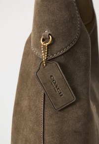 Brown suede shoe with a smooth finish, featuring a gold-tone ring and tag with the label "COACH NEW YORK." Accents include detailed stitching.