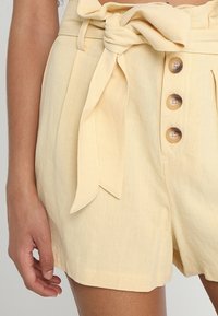 Jasnożółte lniane szorty z wiązaniem w talii, plisami i trzema brązowymi guzikami z przodu jako zapięcie.