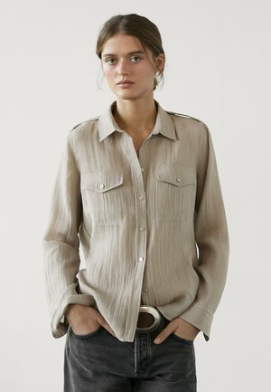 Chemise beige à boutons avec un tissu texturé, deux poches avant et des accents sur les épaules ; portée avec un jean foncé et une grande ceinture.