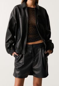 Veste aviateur en cuir noir avec poignets élastiques, portée sur un haut transparent à pois brillants. Assortie à des shorts en cuir noir à texture lisse.