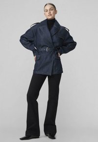 Žena stojaca s rukami v bok, oblečená v tmavomodrej rifľovej bunde so zápästím, čiernom roláku, čiernych zvonových nohaviciach a čiernych špicatých topánkach.