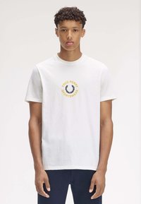 Joven con cabello rizado que lleva una camiseta blanca de Fred Perry Sportswear y pantalones azul marino, de pie contra un fondo simple.