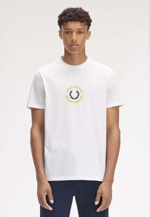 Young man with curly hair wearing a white Fred Perry Sportswear T-shirt and navy pants, standing against a plain background.
