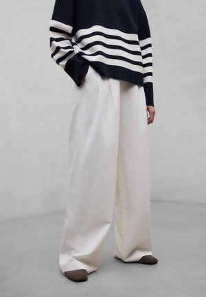 Personne portant un pull bleu marine à rayures blanches, un pantalon blanc à jambes larges et des chaussures tissées foncées, debout devant un fond gris uni.