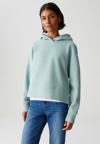 Donna con felpa con cappuccio azzurro chiaro e jeans blu, in piedi contro uno sfondo grigio chiaro uniforme.