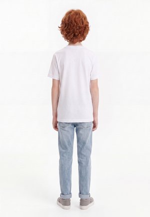 Barn med rødt krøllet hår står med ryggen til iført en ensfarvet hvid t-shirt, lyseblå jeans med opsmøg, og grå sneakers.