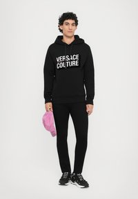 Čierna mikina s kapucňou s logom "Versace Jeans Couture", v kombinácii s čiernymi nohavicami. Drží ružovú šiltovku a má na sebe čierne tenisky.