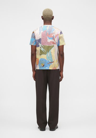 Άτομο με κοντά ξανθά μαλλιά που φοράει πολύχρωμο πουκάμισο με σύντομο μανίκι και αφηρημένο σχέδιο και μαύρο παντελόνι, στέκεται με την πλάτη προς τα εμάς σε απλό φόντο.