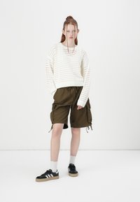 Knit white sweater with textured pattern, paired with olive green cargo shorts featuring side pockets and drawstring. Black sneakers with white stripes.
