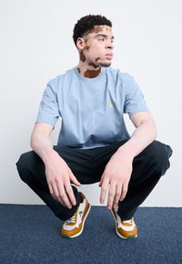 Svetlomodré bavlnené tričko, vo voľnom strihu, s malým žltým logom. Čierne nohavice a hnedé, biele a zelené tenisky dopĺňajú outfit.