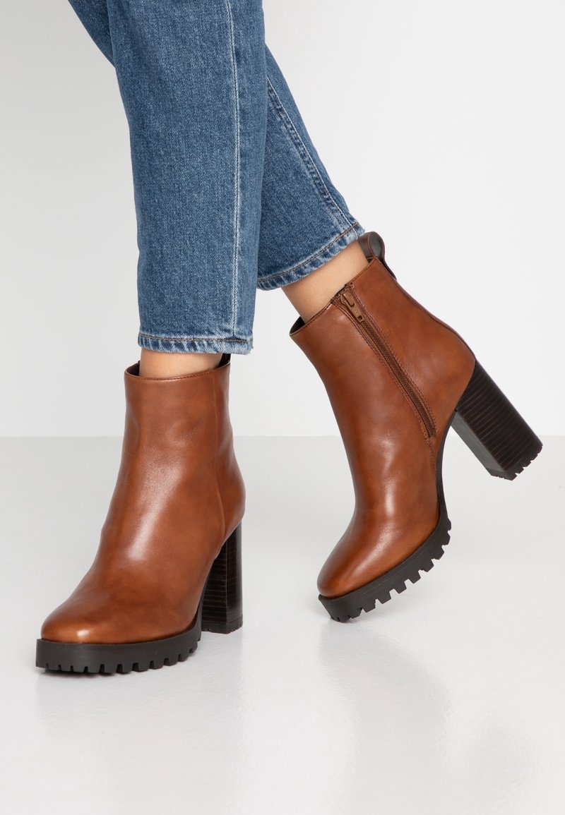 Bottines en cuir marron à talons épais avec fermetures éclair latérales portées avec un jean en denim bleu sur un fond blanc.