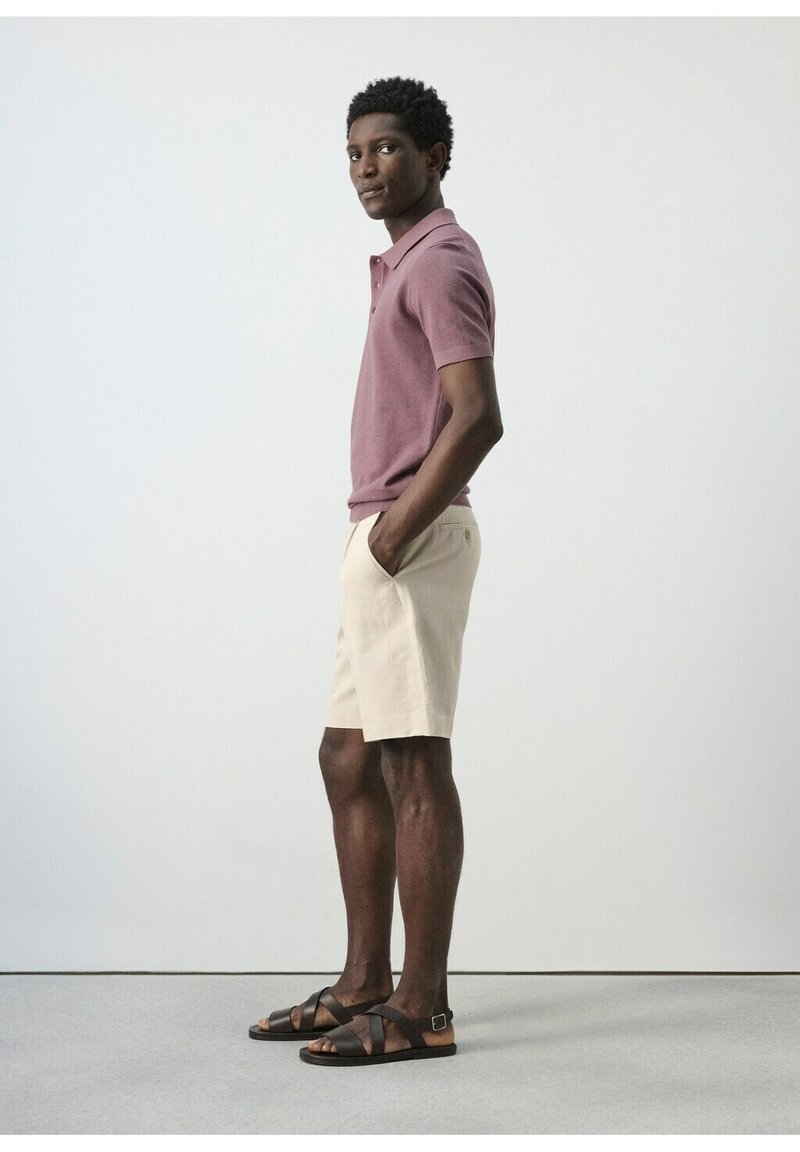 Homme debout de profil portant un polo mauve, un short beige et des sandales marron foncé, les mains dans les poches.